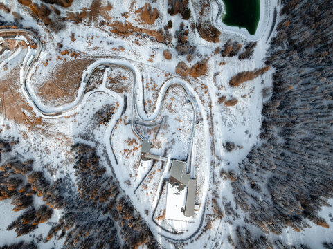Aerial view of the Cesana Pariol bobsleigh track winding through a snow-covered forest landscape with evergreen trees and a small pond in Cesana Torinese, Piedmont, Italy.