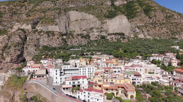 Aerial view of Meta di Sorrento colorful buildings perched on tuff cliffs with coastal road, arched viaduct, and dramatic rocky mountain backdrop, Naples, Italy