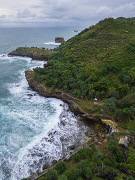 Aerial view of Pantai Jogan, featuring a waterfall cascading onto rocky beach, lush green cliffs, and crashing waves in Kecamatan Tepus, Daerah Istimewa Yogya, Indonesia.