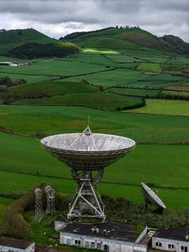 Aerial view of a large satellite dish on Sao Miguel Island surrounded by lush green rolling hills and pastures under a cloudy sky, Azores, Portugal.