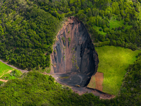 Aerial view of the Tecnovia Acores quarry site showing a deep excavation pit with dark volcanic soil, surrounded by lush green forest and grassy fields in Rabo de Peixe, Acores, Portugal.