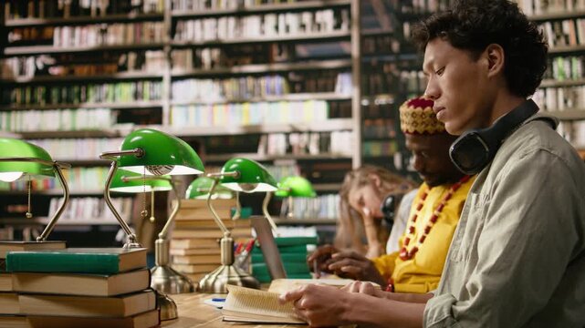 Asian student with headphones reading at library table behind books and green lamps, with another learner studying in blurred background, peers learning in study session, for online learning.