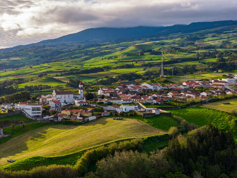 Aerial view of the village of Salga featuring a church with a red roof, surrounded by lush green rolling hills, agricultural fields, and stone walls in Salga, Azores, Portugal.