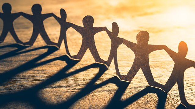 Chain of paper figures holding hands on wooden surface with sunset lighting. Concept of teamwork, community unity, and business partnership.