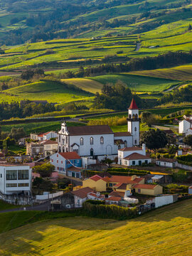 Aerial view of the Church of Salga surrounded by traditional houses and vibrant green rolling hills in the rural landscape of Salga, Azores, Portugal.