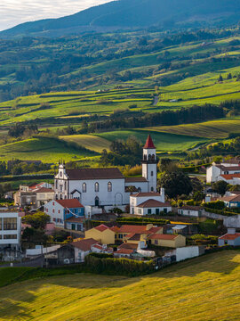 Aerial view of the church with a red-roofed tower nestled among green rolling hills and traditional houses in Salga, Azores, Portugal.