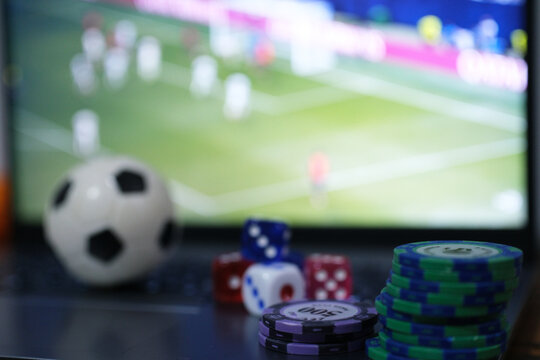 Online soccer gambling. Image of a ball, gambling tokens, and dice in front of a laptop with a live soccer broadcast. Live scores. Gambling bets.
