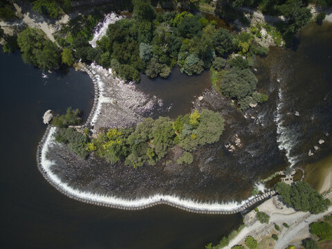 Aerial view of the Tamega River flowing over a historic stone weir surrounded by lush green trees and vegetation in Amarante, Porto District, Portugal.