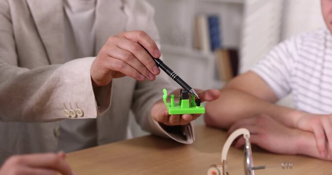 Teacher with DC motor model explaining physics to students at wooden desk in classroom, closeup. Camera moving in