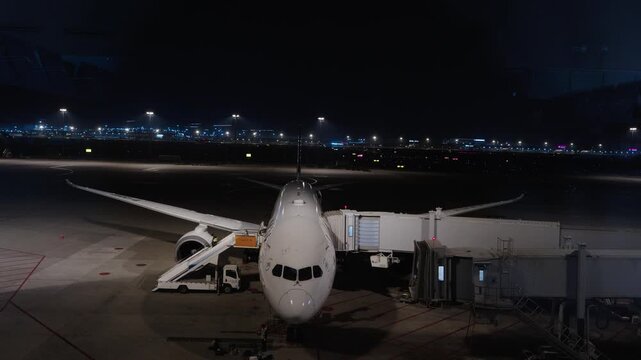 Commercial airplane docked at jet bridge on illuminated airport tarmac at night timelapse