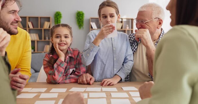Family children grandparents play boardgame with cards. Parents and kids engage at the table at home. Warm expressions show teamwork indoors, boosting memory skills. Clear idea of family learning.