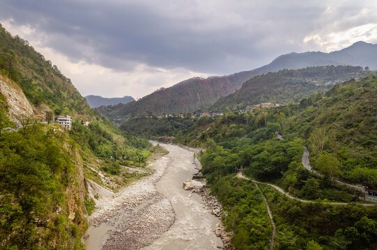 Aerial view of the Alaknanda River flowing through the lush green Himalayan mountains with a winding road and cloudy sky in Chamoli Gopeshwar, Uttarakhand, India.
