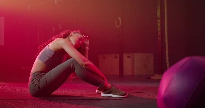 African American woman seated doing crunches, catching breath in gymwear on mat near purple ball