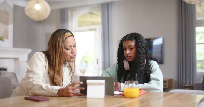 African American mom positioning tablet, helping teenage daughter with homework on papers at table