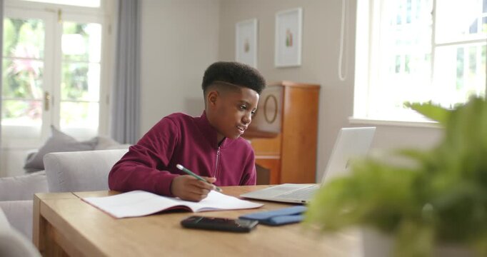 African American male Youth studying at home, reading laptop and writing in notebook with pen