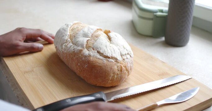 Male hands entering left at counter by window near crusty loaf on board, reaching knife, preparing