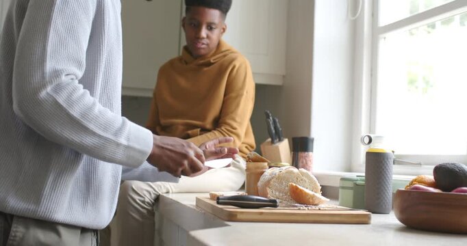 African American man and teen boy preparing snack, opening jar, scooping spread onto roll by window