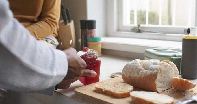 Hand is loosening jam lid and spreading jam on sliced loaf at window counter for breakfast
