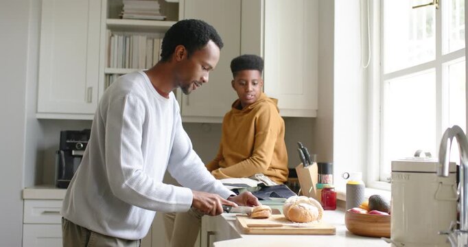 African American dad slicing bread for breakfast in kitchen as teenage son reading, glancing window
