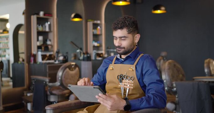 African American male barber sitting in shop tan apron shears replying to tablet checking roster