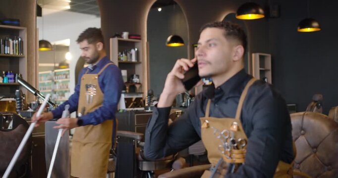 Male barber sitting in shop seat answering phone after alert, in brown apron while helper mopping