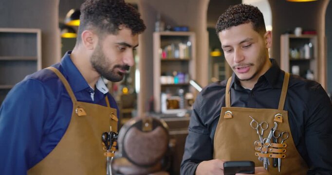 Male barbers in leather aprons at center barbershop, one checking smartphone, other joining to view