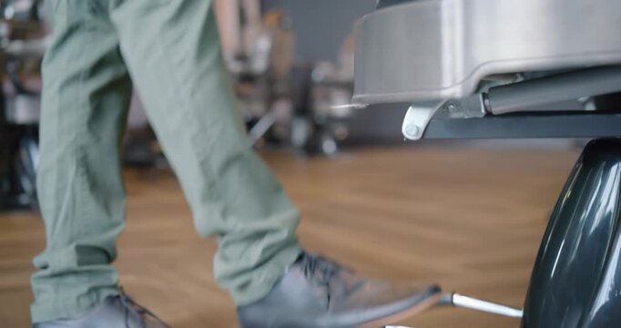 Male barber in apron entering from left in salon, pumping pedal, preparing client chair, copy space