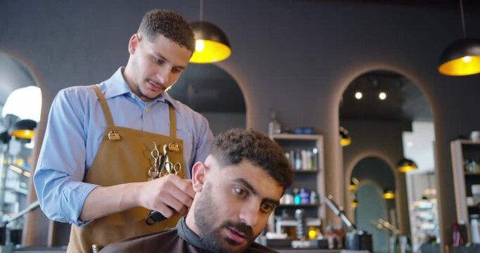 African American male aproned barber cutting client after tilt at shop refining line with clipper