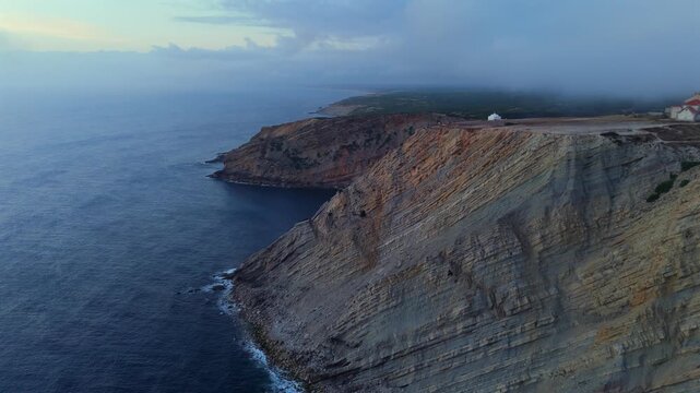 Drone view of steep layered rock cliffs rising above the ocean with textured stone formations along a rugged shore