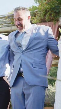 Vertical video: Friend stepping behind nudging groom in suit walking down garden aisle with bouquet