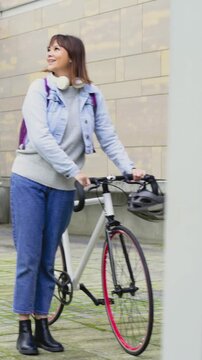 Vertical video: Entering frame, woman in denim jacket pushing bicycle across courtyard, commuting