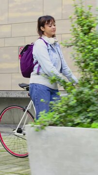 Vertical video: Glancing upward, woman pushing white bike across plaza heading to door with helmet