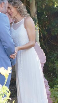 Vertical video: Reaching bride in white gown holding hands, pressing foreheads at wedding arch