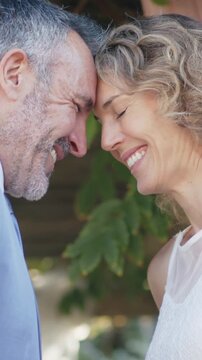 Vertical video: Holding hands, groom and bride in white dress touching foreheads at garden ceremony
