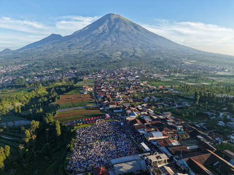 Aerial view of Lapangan Garung with a large crowd gathered at the base of Mount Sumbing, surrounded by rooftops and green fields, Kecamatan Kalikajar, Jawa Tengah, Indonesia.
