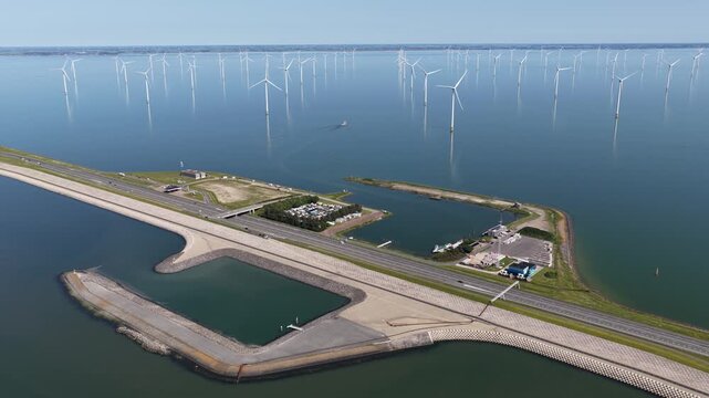 Impressive aerial perspective of an offshore wind farm with numerous turbines standing in calm water, producing clean, renewable energy next to a long causeway with a small harbor