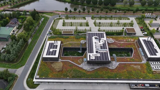Sustainable modern architecture featuring a large sedum extensive green roof with solar panels for renewable energy, seen from a descending aerial perspective showing the nearby parking and canal