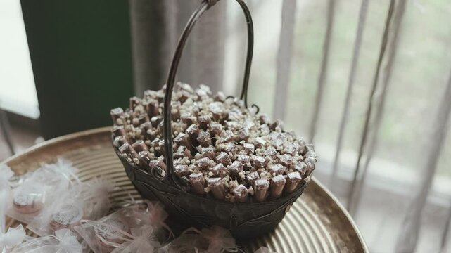 Metal basket filled with small wrapped wedding favor gifts on golden tray with white feathers beside window at reception