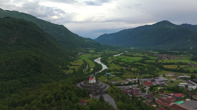 Soča River winds through lush green valley below Kobarid Ossuary WWI memorial church, with forested mountain slopes, farmland and cloudy skies over Kobarid, Slovenia. Aerial drone panorama view