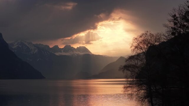 Intense golden sunset rays bursting through storm clouds over alpine lake with snowy peaks and tree silhouette Switzerland