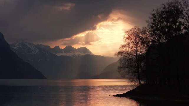 Dramatic sunset light breaks through clouds over a calm lake with silhouetted trees