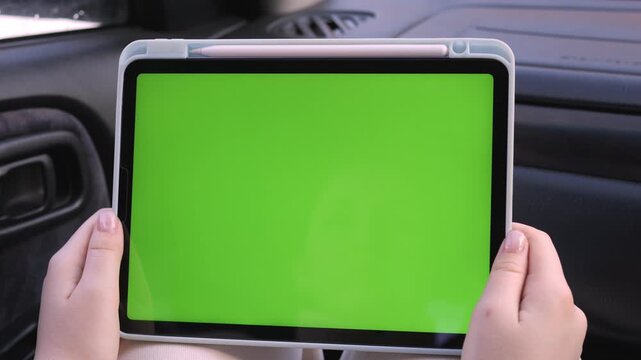Hands hold tablet with green screen inside car. Person appears calm and focused. Ideal for travel, navigation, or digital content overlays. Clear view of device and car interior