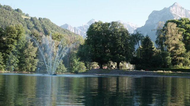 Peaceful view of Walensee Lake in Switzerland with a fountain, reflective water, lush trees, and alpine mountains creating a scenic European landscape.