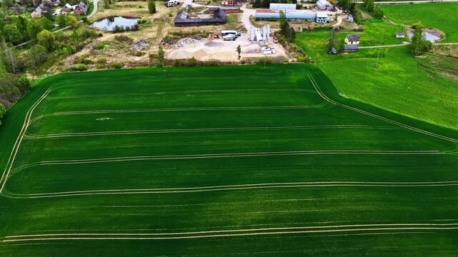 Vibrant green agricultural field with parallel tractor tracks bordering a rural industrial area