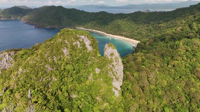 Stunning high-angle aerial drone shot of a secluded sandy crescent beach hemmed in by lush jungle-covered mountains and steep karst limestone cliffs along the scenic coast of Palawan, Philippines.