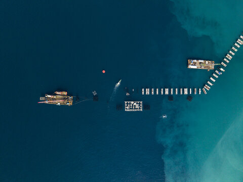 Aerial view of the Oil Fuel Terminal Port construction site with a jetty extending into the sea, construction barges, and a boat in the turquoise waters in Manggis, Bali, Indonesia.