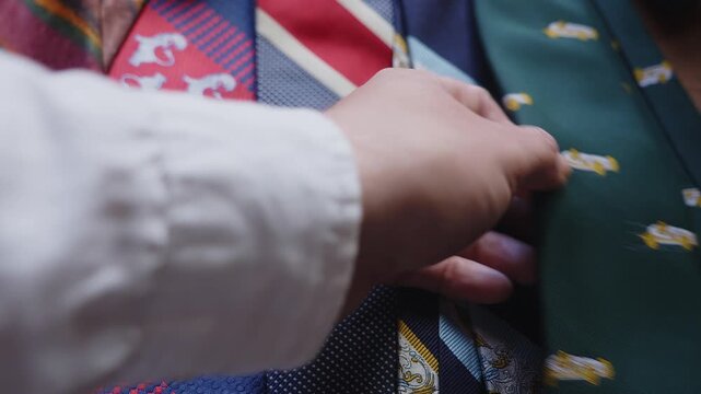 A person compares multiple neckties while deciding on a final look. The scene suggests personal style, fashion choices, preparation, identity, and vintage menswear aesthetics.