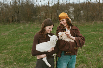 Obraz premium Two women, friends, outdoors in a grassy field cuddle goat kids, sharing smiles. A lifestyle moment with animals and rural scenery, highlighting care, connection, and gentle play in a countryside