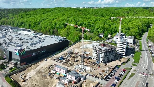 Cinematic aerial shot of a busy construction site with cranes and new building development next to a shopping mall and lush green hills in Gdynia.