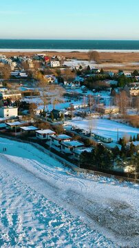 A cinematic birds eye view of a quiet seaside village covered in snow during a bright winter day. High quality drone shot of residential houses and the Baltic sea horizon.